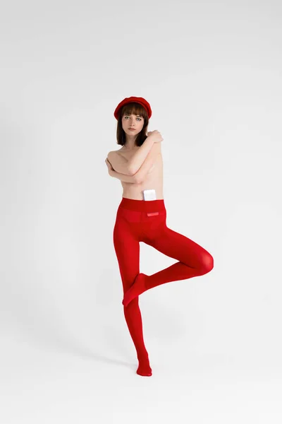 Stock image Young beautiful girl only in red tight and a beret, hands covering her bare chest. Sensual slim girl posing topless. Isolated on white background. Full length, vertical.