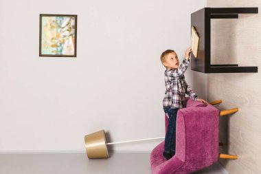 Küçük çocuk baş aşağı odada, elini masaya uzatıp kitap okuyor. Odanın duvarında duran çocuk. Ters çevrilmiş oda.