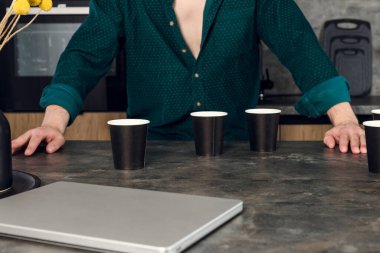 Kahve bağımlısı uykucu adam mutfakta içmeyi bırakamıyor, bir sürü fincan masanın etrafına saçılmış. Kafein bağımlılığı kavramı.