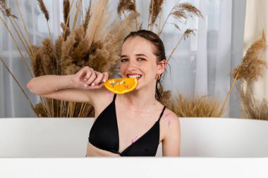 Çekici bir genç kız banyoda oturmuş elinde çiçek yaprakları ve portakal dilimleriyle poz veriyor. Güzellik ve cilt bakımı.