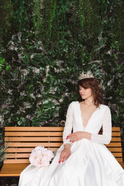 Zarif beyaz elbiseler içinde güzel bir gelin ve taç çiçeklerin önündeki ahşap bir bankta ve çiçekli bir kemerde oturuyor. Bahçedeki gelin..
