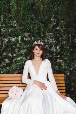 Zarif beyaz elbiseler içinde güzel bir gelin ve taç çiçeklerin önündeki ahşap bir bankta ve çiçekli bir kemerde oturuyor. Bahçedeki gelin..