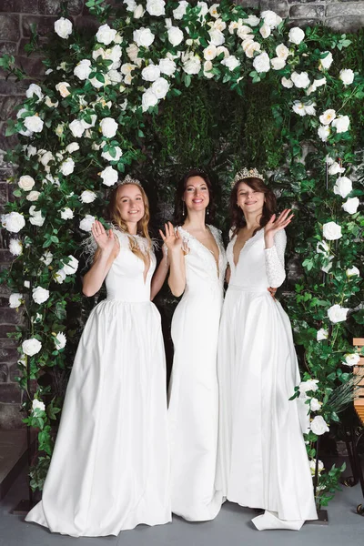 stock image Three beautiful brides in elegant white dresses in front of plants and an arch with flowers. Group of brides in a garden.