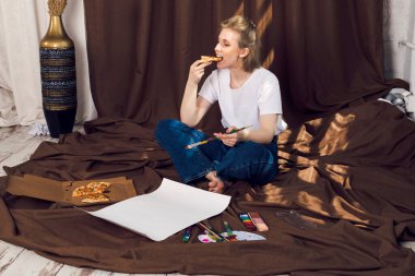 Şirin, rahat sarışın kız, ressam ya da sanatçı rahat bir atölyede kahverengi bir bez üzerinde uzanmış pizza yiyor ve güneş ışınlarının altında resim yapıyor..