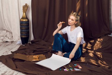 Şirin, rahat sarışın kız, ressam ya da sanatçı rahat bir atölyede kahverengi bir bez üzerinde uzanmış pizza yiyor ve güneş ışınlarının altında resim yapıyor..