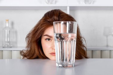 Bir kız bir bardak suyun içinden bakıyor, yüzünü bozuyor. Ürün vitaminleri, hidratlaştırıcı içecekler, alkol.