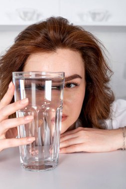 Bir kız bir bardak suyun içinden bakıyor, yüzünü bozuyor. Ürün vitaminleri, hidratlaştırıcı içecekler, alkol.