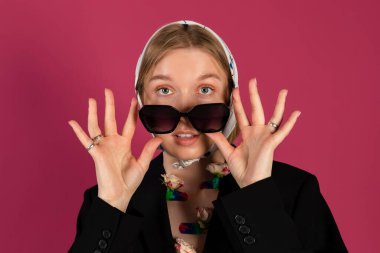 Güzel, narin, genç, sarışın bir kızın portresi şık bir siyah ceket ve göğsüne takılı yaratıcı çiçekler ve başörtüsü takıyor. Pembe arka planda izole edilmiş.