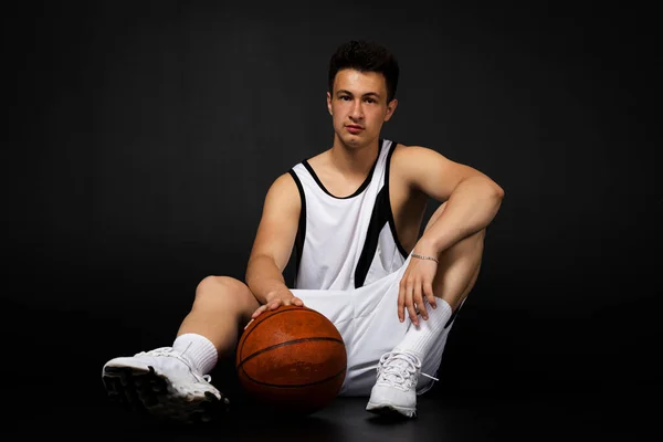 Przystojny Młody Koszykarz Białej Odzieży Sportowej Siedzi Podłodze Pozuje Piłką — Zdjęcie stockowe