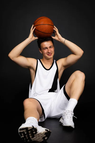 身穿白色运动服的年轻漂亮的篮球运动员坐在地板上 摆姿势带球 被隔离在黑色背景下 — 图库照片