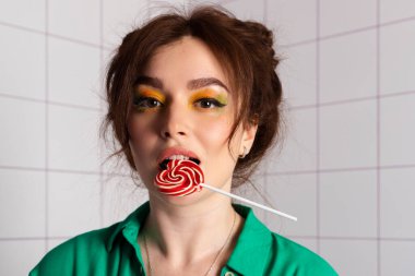 Çekici, güzel bir kız stili. Parlak makyajı, çift katlı saçı, duygusal ifadesi, banyoda poz vermesi, kalp şeklinde lolipop yemesi, temiz minimalist tarzı, beyaz fayanslar.