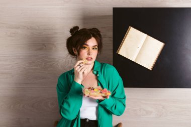 Çekici, güzel bir kız. Parlak makyajlı, çift katlı çörek saçlı, duygusal, yüz ifadeli, bir sürü tatlı yiyen, masa ve kitaplı ters oda arkaplanı.