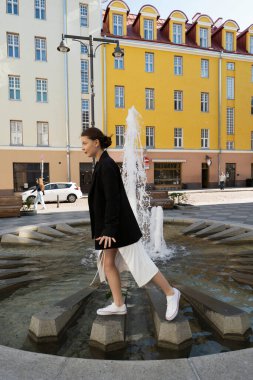 Beyaz elbiseli ve siyah ceketli güzel bir esmer bir Avrupa şehrinin sokaklarında bir çeşmeye ve süslü eski binalara karşı yürüyor.