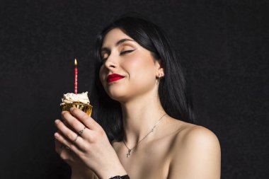 18. yaş gününü kutlayan siyah saçlı ve elbiseli zarif giyimli genç bir kız portresi. İçinde mum olan küçük bir pasta tutuyor. Siyah arkaplanda izole.