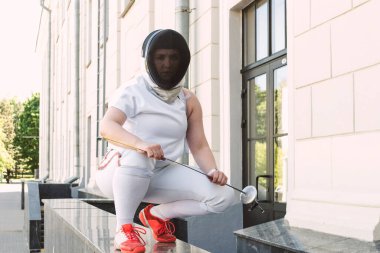 Kadın eskrim sporcusu, maske takıyor, beyaz eskrim kıyafeti giyiyor ve kılıcı tutuyor, dışarıda poz veriyor.