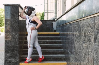 Kadın eskrim sporcusu, maske takıyor, beyaz eskrim kıyafeti giyiyor ve kılıcı tutuyor, dışarıda poz veriyor.