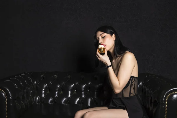 肖像18歳の誕生日を祝う黒い髪とドレスを持つエレガントな服を着た若い美しい女の子 黒い革のソファに座って小さなケーキを食べる 黒の背景 — ストック写真