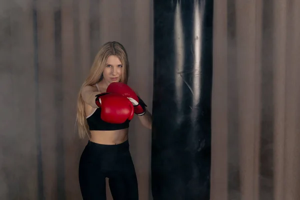タイトな黒のスポーツウェアとボクシングの手袋を身に着けている魅力的なスポーティーな女の子 パンチバッグでトレーニング 煙とジムでポーズ — ストック写真