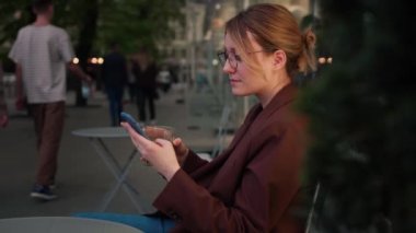 Dışarıda cep telefonuyla yazan mutlu genç bir kadın. Gözlüklü neşeli beyaz kız şehir arka planında akıllı telefon kullanıyor. Sosyal medya, kurye, mafya uygulaması, video izleme, online alışveriş