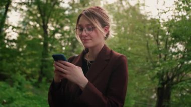 Parkta telefon kullanan bir kadın. Dışarıda elinde cep telefonu olan beyaz bir kız. Elinde akıllı telefon tutan ve gülen yüzüyle ekranda yazan bir kadın. Yüksek kalite 4k görüntü