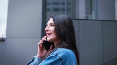 Kenar manzaralı bir kadın şehir caddesinde yürüyen bulutlu bir günü arıyor. Mavi ceketli mutlu, gülen kız dışarıda akıllı telefondan konuşuyor. Neşeli bayan gezici sohbetin keyfini çıkarın. Yüksek