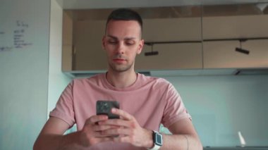 Yakışıklı, beyaz, profesyonel, elinde modern akıllı telefondan mesaj tutan biri. Genç işadamı mutfakta oturan mobil uygulamaları kullanıyor. Yüksek kalite 4k görüntü