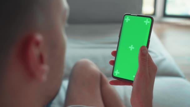 自宅で若い白人男性緑のモックアップ画面でスワイプスマートフォンを使用してソファの上に横たわる スワイプを行う ジェスチャーをスクロール 携帯電話を使用して男 インターネットソーシャルネットワークブラウジング — ストック動画
