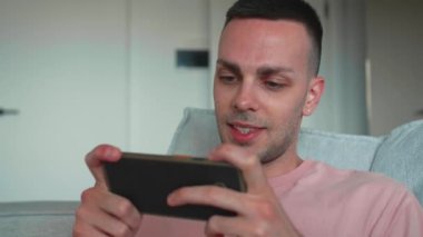 Telefon, evinin oturma odasındaki kanepede bir adamı mobil oyun oynaması için oynamak. İnternet, teknoloji ve bilgisayar oyunları bir evde rahatlamak için internetten oynayan bir erkek oyuncu ineği. Yüksek kalite 4k