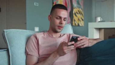 Kötü haber, cihaz sorunu, uygulama çökmesi konsepti. Genç adam oturma odasında kanepede oturup elinde akıllı telefon tutarak olumsuz mesajlar okuyor, rahatsız oluyor, alet sorunu yaşıyor, kablosuz ağ bağlantısını kaybediyor, e-posta yolluyor.