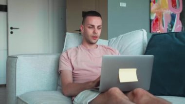 Oturma odasındaki işi bittikten sonra kanepeye oturmuş laptopu kapatan beyaz bir adam. Genç adam internette çalışıyor, uzaktan kumandalı kanepe. Defter bilgisayarında e-öğrenme, internette gezinme