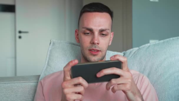 穿着粉色衬衫的外向的高加索人正在沙发上的客厅里玩手机 快乐年轻英俊的小伙子坐在新家的沙发上 高质量的4K镜头 — 图库视频影像