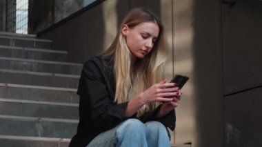 Mutlu beyaz kadın portresi, binanın yanında oturuyor, akıllı telefon kullanıyor. Teknoloji konsepti. Yaz. Yüksek kalite 4k görüntü