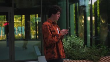 Modern ofis binası yakınlarındaki caddede yürüyen gülümseyen adam elinde telefonla mutlu internet haberleri tutarak akıllı telefon kullanarak e-posta yazma mesajlarını sosyal ağda kontrol ederek mobil uygulama kullanıyor.