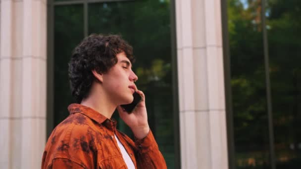 穿着橙色衬衫的年轻人一边讲着手机 一边走在大街上现代建筑商业办公室的背景下 高质量的4K镜头 — 图库视频影像