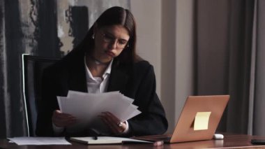 Bir kadın bir odada bir masada oturuyor, elinde kağıtlar ve dizüstü bilgisayar kullanıyor. Gözlük takıyor ve bir ofis ortamında beyaz bir işçiye benziyor.