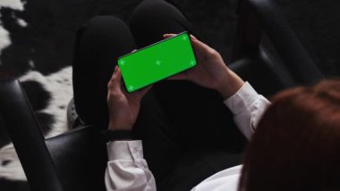 Yakın plan kadın elleri, sandalyede oturan yeşil ekranlı telefondan izliyor. Kadın tarafından kullanılan yeşil ekran cromakey telefon. Teknoloji yaşam tarzı. Yüksek kalite 4k görüntü