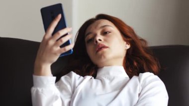 Üzgün, tatminsiz, genç bir kadın evinde kanepede oturup cep telefonu tutarak vakit geçiriyor. Kötü ruh hali yaşam tarzı konsepti. Yüksek kalite 4k görüntü