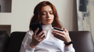 Evdeki koltukta oturan, elinde kredi kartı ve akıllı telefon tutan gülümseyen genç bayan müşteri. İnternetten alışveriş yapan mutlu bayan alışverişçi kolay taşınabilir ödemeler yapıyor. E-bankacılık uygulama servisi