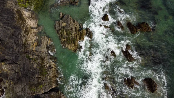 Waves break on the rocky shore of the Celtic Sea, top view. White foam on the sea waves. The coastline of the Atlantic. Turquoise sea water. Breathtaking seaside landscape. Aerial photo.