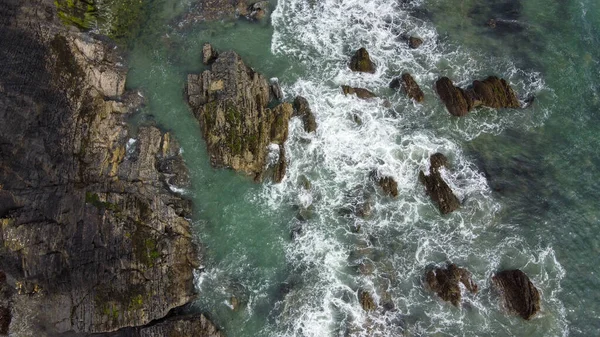Waves break on the rocky shore of the Celtic Sea, top view. White foam on the sea waves. The coastline of the Atlantic. Turquoise sea water. Breathtaking seaside landscape. Drone photo.