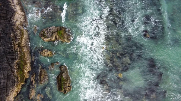 Waves break on the rocky shore of the Celtic Sea, top view. White foam on the waves. The coastline of the Atlantic. Turquoise sea water. Breathtaking seaside landscape. Drone photo. View from above.