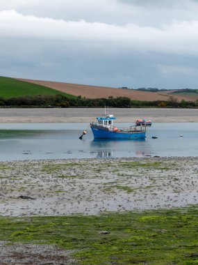 Dalgalar çekildiğinde sığ sularda bir tekne. Bulutlu bir gökyüzünün altındaki tepeler. Çamur ve yosun. Peyzaj.