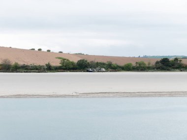 Kumlu deniz kıyısı bulutlu bir günde. Sakin su yüzeyi. Kıyıdaki eski bir geminin kalıntıları. Tarlaya sürülmüş. Minimalist manzara.