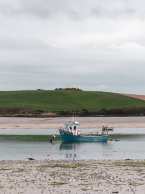Küçük eski bir balıkçı teknesi, bulutlu bir günde, sığ sularda demir atar. Bulutlu bir gökyüzünün altındaki tepeler. Çamur ve yosun. Peyzaj.