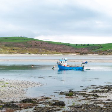 Küçük eski bir balıkçı teknesi, bulutlu bir günde, sığ sularda demir atar. Gökyüzünün altındaki tepeler.