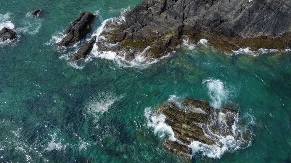 Black moss-covered rocks among turquoise waves of the Celtic Sea. White sea foam on the waves. Waters of the Atlantic Ocean. Drone point of view.