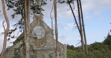 Tall coniferous trees in front of a nineteenth-century stone church in Darrara. Christian church on a cloudy evening. Sacred Heart Roman Catholic Church (built in 1897).