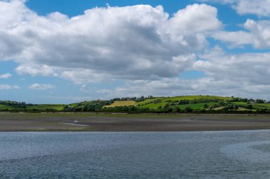 White cumulus clouds in the sky over the hilly Irish countryside. Green hills on a nice summer day. Irish countryside, County Cork. Green grass field near body of water under white clouds and blue sky