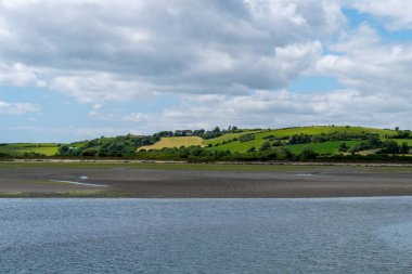 White clouds in the sky over the hilly Irish countryside. Green hills on a nice summer day. Irish countryside, County Cork. Green grass field near body of water under white clouds and blue sky