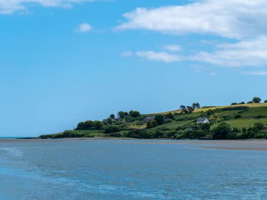 Beautiful clear sky over a calm water surface on a summer day. The picturesque green coast of Ireland. Several buildings on the hill. Seascape. Green trees